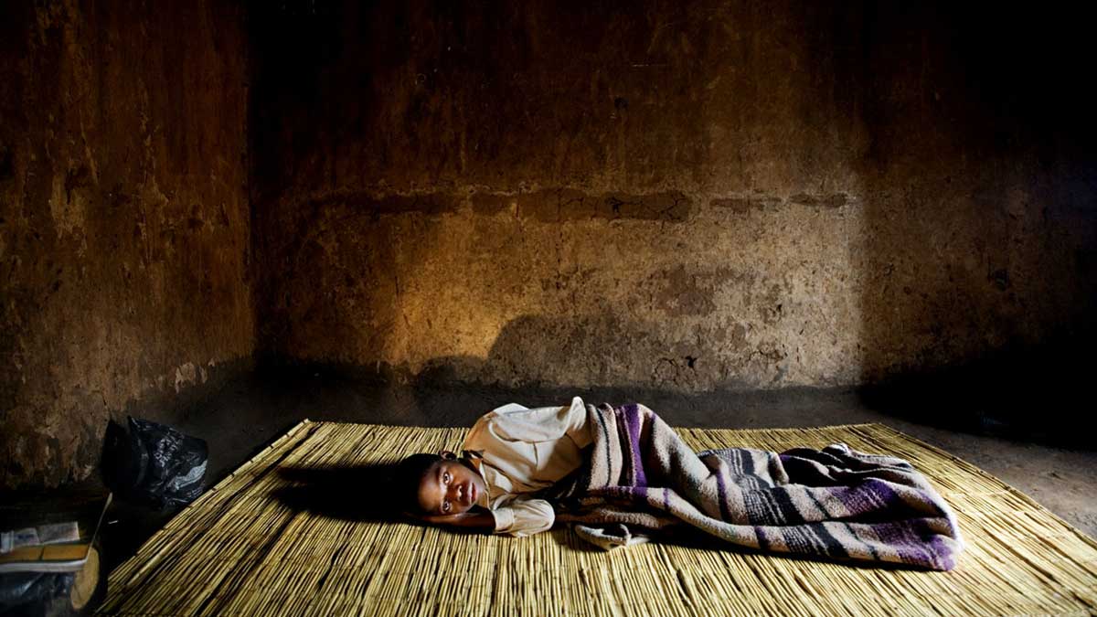 Photo of a person lying in a house on a straw mat covered by a blanket.